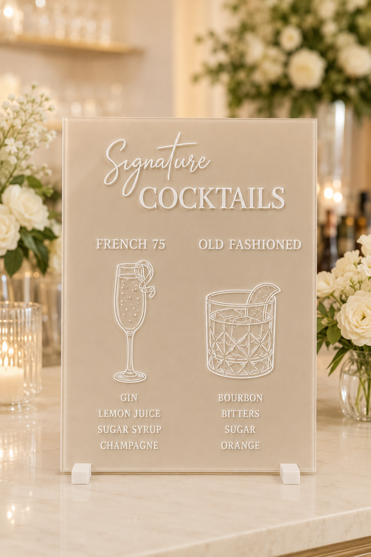 Personalised champagne acrylic cocktail sign with white lettering, displayed on a wedding bar table with floral decorations.